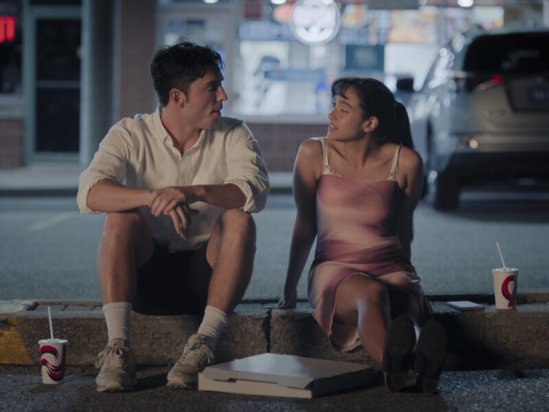  Dois jovens em um momento de intimidade sentados no meio-fio de um estacionamento à noite. O rapaz, Benny, à esquerda, veste uma camisa branca de botão, shorts escuros e tênis. Ele se inclina para a frente com os braços sobre os joelhos, olhando para a jovem. A jovem, Carmen, à direita, com cabelo escuro preso, veste um vestido rosa de alças e olha de volta para ele. Entre eles, no chão, há uma caixa de pizza branca e copos com canudo, sugerindo um encontro casual. Ao fundo, um carro está estacionado e luzes desfocadas indicam um ambiente urbano.