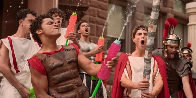 A imagem captura um momento de alta energia com um grupo de jovens em fantasias de romanos em frente a um prédio de tijolos. A cena é caótica e festiva. Em destaque, Benny, um rapaz de armadura de couro e túnica vermelha reage com o rosto para o alto enquanto água é esguichada nele de pistolas de brinquedo. À sua direita, outro rapaz, fantasiado de imperador com uma toga, capa vermelha e coroa de louros, grita em êxtase, segurando um grande bastão. Ao fundo, outros jovens em trajes semelhantes participam da bagunça, um deles também gritando e segurando uma pistola de água. A cena sugere um trote de faculdade ou uma festa temática.