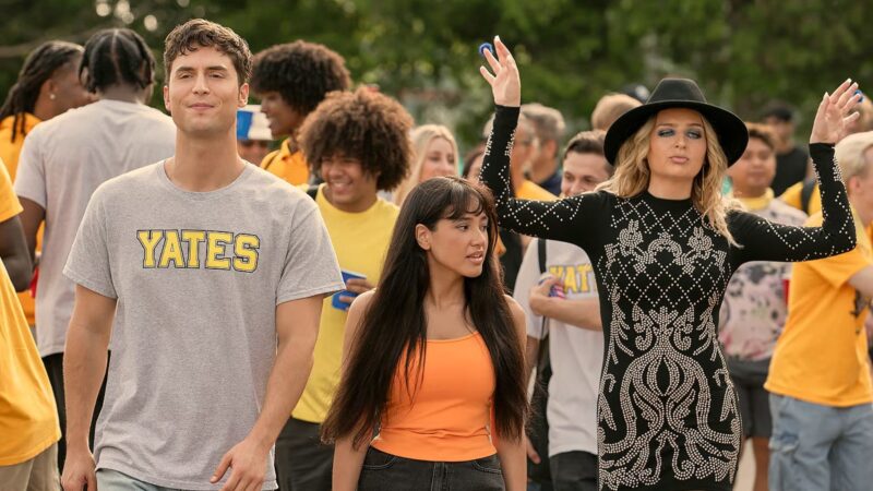 Em uma cena externa, três jovens estão em primeiro plano com uma multidão ao fundo. À esquerda, Benny, um jovem branco de cabelo castanho sorri, vestindo uma camiseta cinza com a palavra "YATES" estampada em amarelo e azul. No centro, Carmen, uma jovem de cabelos longos e escuros e franja, vestindo uma regata laranja, olha para o lado com uma expressão de preocupação. À direita, Hailee, uma jovem loira e expressiva usa um chapéu preto e um vestido preto de manga comprida com um padrão prateado brilhante, posando com os braços levantados. O fundo está cheio de outros jovens, muitos usando camisetas amarelas.