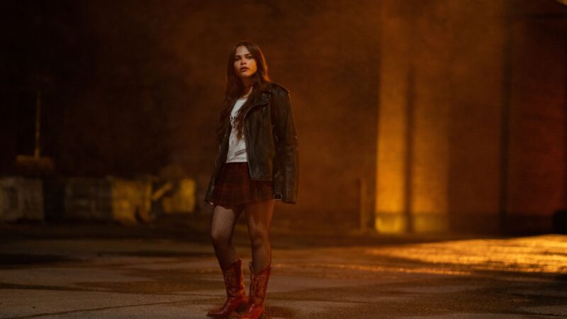 Cena do filme Rua do Medo: Rainha do Baile Na imagem, a personagem Christy encara o seu redor em um galpão. O cenário é noturno e a iluminação amarelada, ao fundo, há paredes e lixo no canto esquerdo. Christy é uma garota na faixa dos 18 anos, de cabelos longos escuros e veste uma bota vermelha, jaqueta de couro preta, camiseta branca, saia xadrez vermelha e meia-calça. 