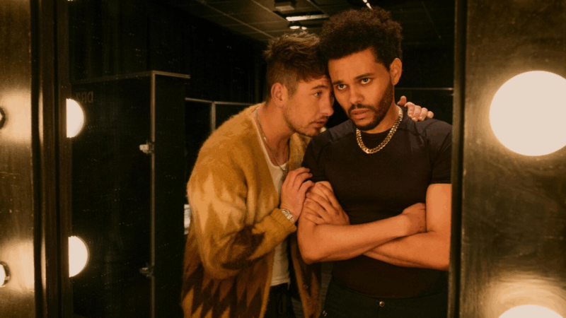 A imagem mostra o ator Barry Keoghan (loiro, usando um casaco de lã claro) com a cabeça apoiada no ombro de Abel Tesfaye (The Weeknd) (cabelo escuro, barba e corrente dourada, com os braços cruzados e olhos marejados) em um camarim com espelhos e luzes.