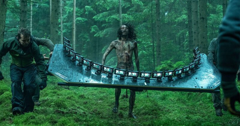 Cena dos bastidores de Extermínio: A EvoluçãoNo centro da imagem, um ator caracterizado como infectado aparece com o corpo coberto de sangue e lama, cercado por uma estrutura semicircular equipada com vários iPhones 15 Pro Max, usados para capturar imagens em 180 graus. Técnicos de produção ajustam o equipamento enquanto capturam a cena de perseguição em uma floresta sombria, cercada por árvores altas.