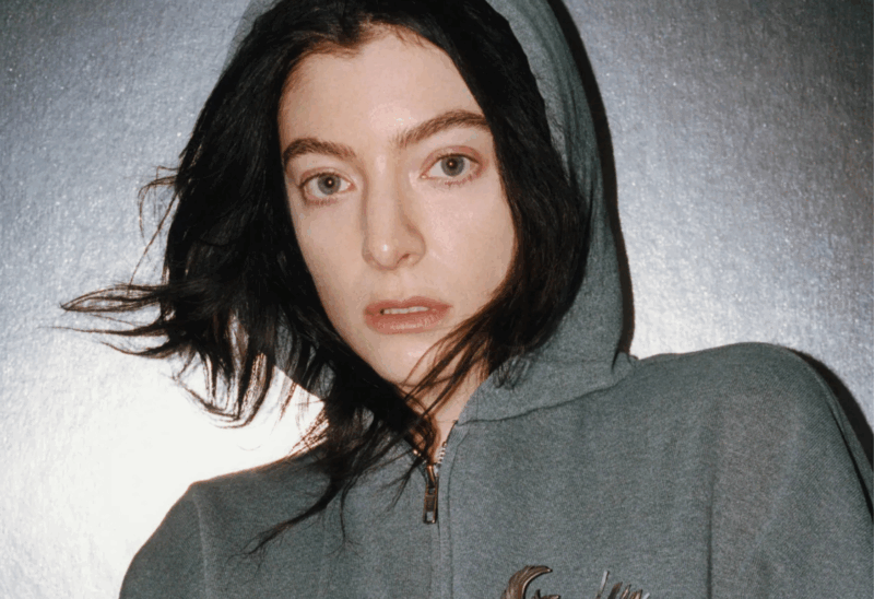 Lorde, mulher branca, aparece em close-up, vestindo um moletom cinza com capuz puxado sobre a cabeça. Seu cabelo preto está levemente bagunçado pelo vento e seus olhos encaram diretamente a câmera, transmitindo uma expressão séria e vulnerável. O fundo metálico e prateado cria um contraste frio com o tom da sua pele clara, reforçando a estética crua e intimista associada à era Virgin.