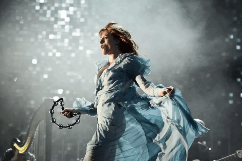 Foto de um show da banda Florence + The Machine. Nela, vemos Florence Welch, uma mulher branca de cabelos ruivos. Ela veste um vestido longo azul. Ela está correndo, de forma com que o vestido, ao mesmo tempo que esvoaça, também cola em seu corpo. Ela também segura uma meia lua com sua mão direita. Ao fundo, luzes de um palco