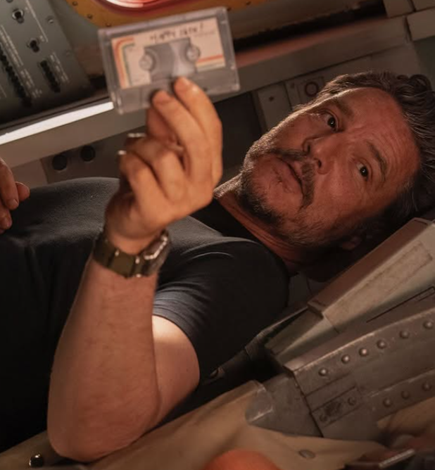 Pedro Pascal, um homem chileno-americano com cabelos curtos, caracterizado de Joel, veste uma camiseta preta e usa um relógio no pulso esquerdo. Na mão esquerda, segura uma fita cassete pequena preta com adesivo branco e detalhes de arco-íris. O personagem se encontra deitado em um assento enquanto olha para o lado direito e oferece o objeto. O cenário é metálico, como uma cápsula.