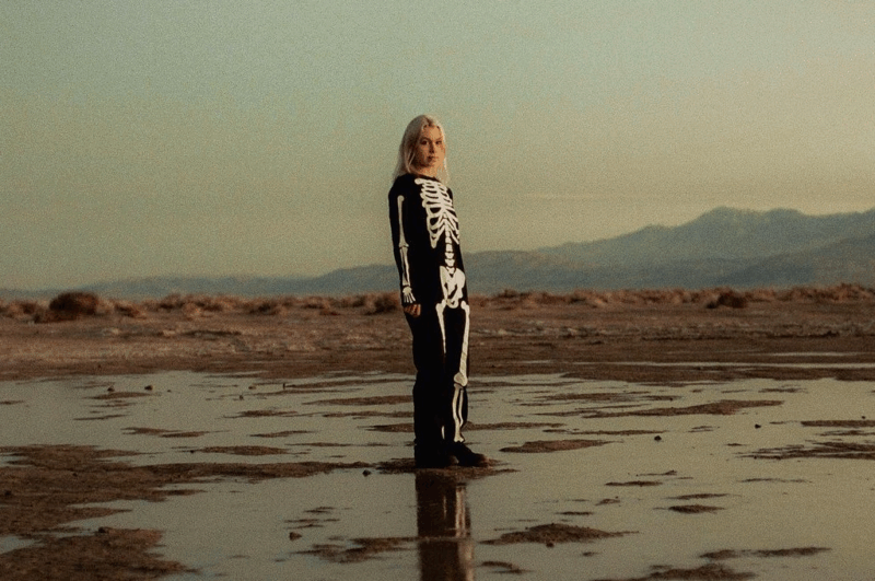 Foto de Phoebe Bridgers, Nela vemos a cantora com um macacão preto com estampa de esqueleto. Ela está em uma planice de terra, com várias poças em volta, enquanto encara a câmera.