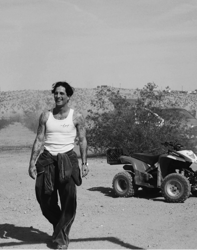 A imagem está em preto, branco e tons de cinza. No lado esquerdo está Damiano David sorrindo com um cigarro na mão abaixada. No lado direito ao fundo, está um quadriciclo, uma árvore e atrás dela um carro. Ao fundo há elevações na terra.