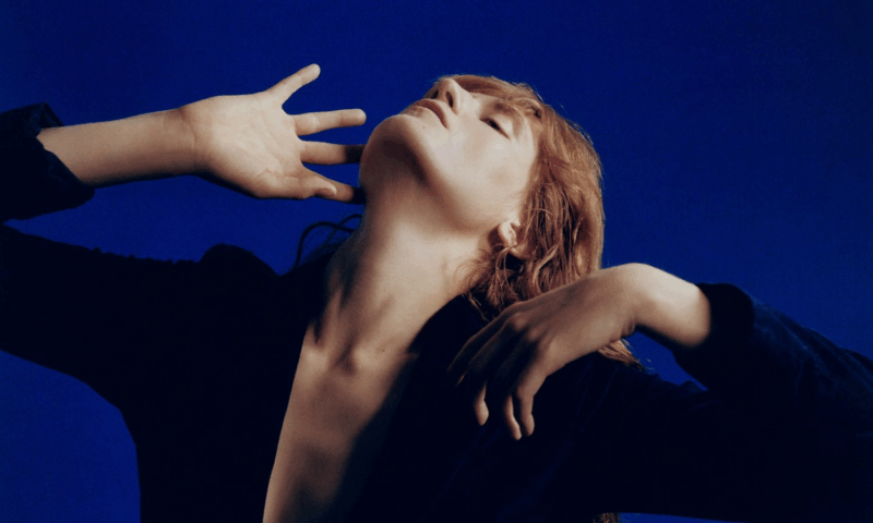 Foto de Florence Welch, vocalista do Florence + the Machine. Ela é uma mulher branca de cabelos ruivos longos. Florence veste uma camisa preta com um decote marcado que vai até a barriga. Ela está com a cabeça arcada para trás e fazendo gestos com suas duas mãos. Ao fundo, uma parede azul