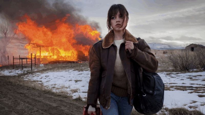 A imagem mostra a atriz Jenna Ortega em um cenário de inverno com neve no chão, vestindo uma jaqueta marrom com gola de carneiro e jeans, enquanto carrega uma bolsa preta e um galão de gasolina vermelho. Ao fundo, uma estrutura em chamas produzindo muita fumaça, e montanhas distantes.