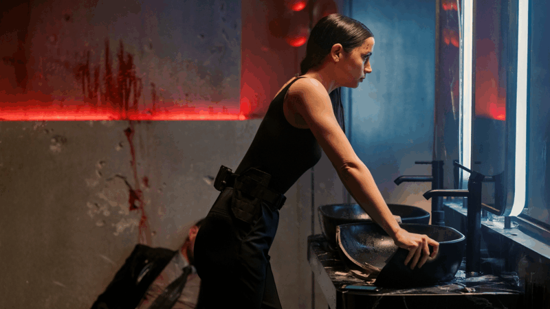 Cena do filme BailarinaNa imagem, Eve está em um banheiro de boate. Há manchas de sangue espalhadas na parede ao lado junto ao corpo de um homem morto, sugerindo que ela deixou um rastro de caos. Diante do espelho, ela se encara intensamente, como se buscasse recuperar as forças antes de seguir com a matança. A personagem veste uma regata preta, calça da mesma cor e um coldre, compondo um visual tático.