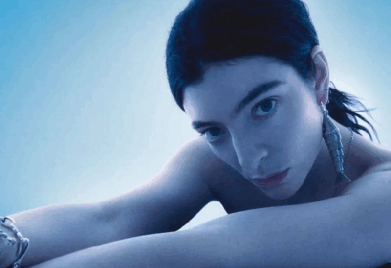 Lorde, mulher branca, posa parcialmente despida contra um fundo azul suave e etéreo. Seu cabelo escuro está preso em um rabo de cavalo baixo, e ela usa um brinco longo e prateado em formato de espiral. Com os braços dobrados à frente do corpo e os olhos fixos na câmera, sua expressão transmite introspecção, força e uma certa melancolia. A iluminação azulada acentua a atmosfera sensorial e aquática que permeia o conceito visual de Virgin.
