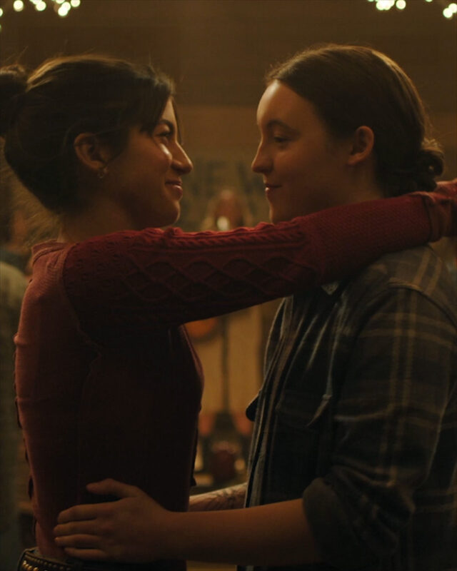 Ao lado esquerdo da imagem, Isabela Merced, uma mulher peruana-americana com cabelos morenos presos em um coque e olhos castanhos, veste uma blusa de lã vermelha enquanto coloca os braços ao redor do pescoço de uma mulher branca, de maneira sensível e delicada, e estampa uma expressão de felicidade, acompanhada dos lábios cerrados em um sorriso largo. Já ao lado direito da composição, há Bella Ramsey que, com os braços de Isabela Merced ao redor do pescoço como em um abraço, olha para a parceira com um sorriso pequeno no rosto. A atriz de Ellie veste uma camisa xadrez azul e branca e permanece com os cabelos presos em um coque baixo. 