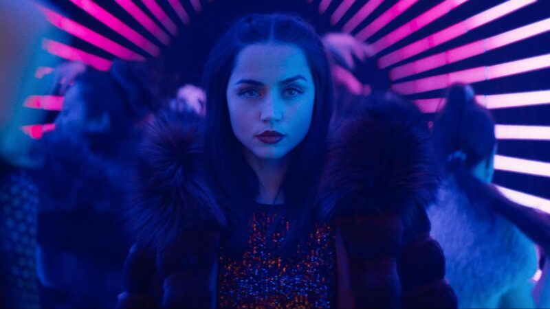 Cena do filme BailarinaNa imagem, a personagem Eve Macarro, interpretada por Ana de Armas, está centralizada em um plano médio com enquadramento frontal. Ela se encontra em um corredor iluminado por luzes de néon rosa e roxa, que formam linhas paralelas ao fundo. Há pessoas ao redor, criando um efeito visual que remete a uma festa em uma boate. Eve é uma mulher branca, magra, de cabelos pretos, e está vestindo um casaco com gola de pelos e uma roupa brilhante em tons de vermelho.