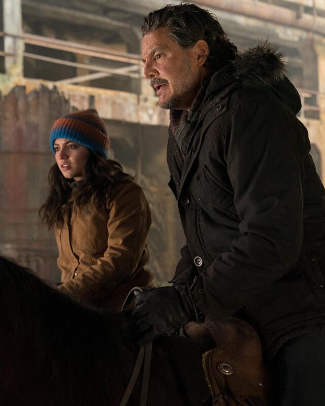 Ao lado esquerdo da imagem, Isabela Merced, interpreta Dina. Ela possui cabelos morenos soltos e uma touca nas cores azul vivo e laranja queimado. Ao lado esquerdo, com os cabelos grisalhos e jaqueta de frio com pelos no gorro, Pedro Pascal interpreta Joel. Ambos os personagens estampam feições preocupadas, com sobrancelhas franzidas e bocas entreabertas tensas. 