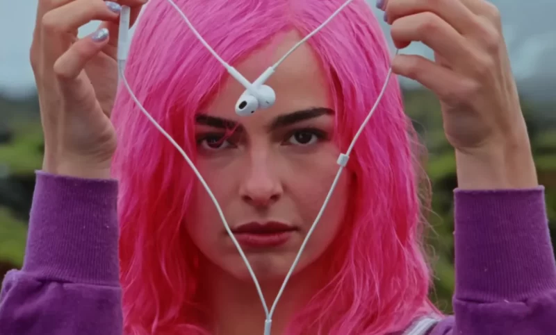 Close-up do rosto de Addison Rae, que tem cabelos rosa choque e uma expressão séria. Ela segura fones de ouvido brancos à sua frente, formando uma forma triangular ou de coração, com os fios emaranhados. Suas unhas estão pintadas de um tom metálico. Ela veste uma blusa roxa, e o fundo é um borrão esverdeado e cinza, sugerindo um ambiente externo.