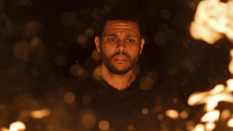 A imagem mostra o artista musical Abel Tesfaye (The Weeknd) com uma expressão aflita, em meio a partículas de chamas e faíscas em um cenário escuro.
