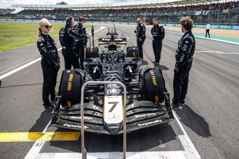 Fotografia do carro, preto com desenhos em fitas também pretas e pintura dourada nas suas extremidades e seus pneus cobertos pelas capas pretas e identificação em amarelo, nas gravações do filme F1, estacionado em sua posição de largada enquanto seis engenheiros da equipe, vestidos em macacões com as mesmas cores, se posicionam nas laterais do veículo em meio a uma pista cinza desgastada e ao fundo arquibancadas para os fãs do esporte. 