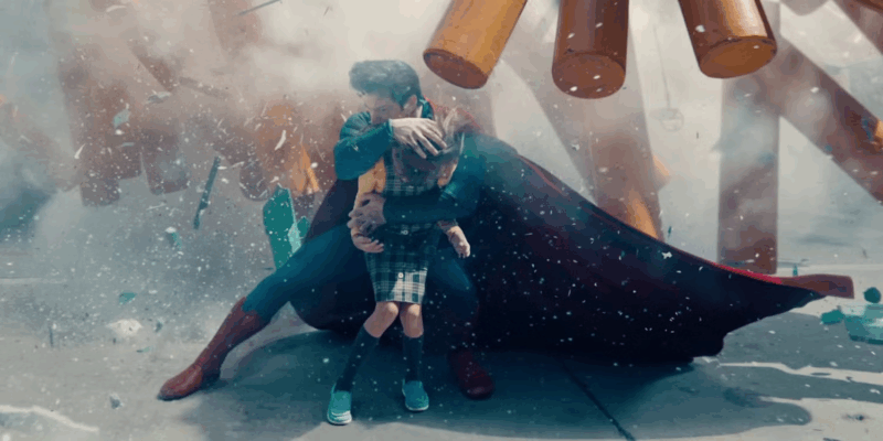 Cena de Superman. Centralizado na imagem está Superman abraçando uma criança. Ele está todo uniformizado, enquanto a criança veste uma camisa amarela e por cima dela uma peça de roupa xadrez verde e branca. Superman está protegendo a criança de destroços amarelos e verdes que estão ao fundo da imagem. 