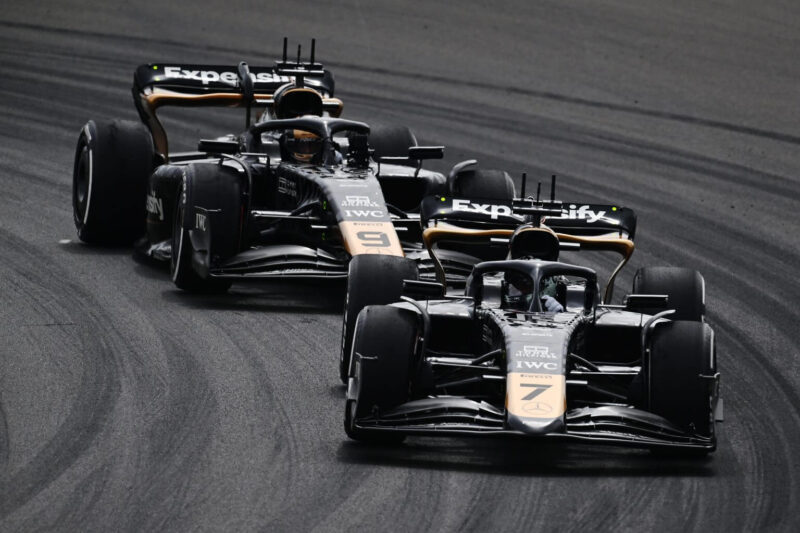 Fotografia dos dois carros de Fórmula 1, pretos por toda sua extensão com exceção do nariz da asa frontal e a aresta inferior da asa que estão destacadas em dourado, enquanto seus patrocinadores estão espalhados estrategicamente pelo automóvel em branco, nos lugares mais analisados pela audiência, em uma cena do filme, ambientada em uma pista de corrida cinza escuro com marcas de pneus em preto.