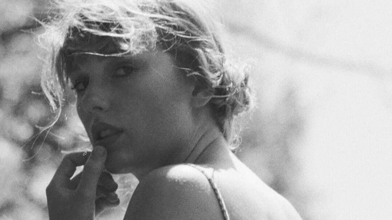 Imagem em preto e branco, em plano fechado, com fundo desfocado de natureza banhada por luz suave. Taylor Swift aparece em destaque, olhando sobre o ombro em direção à câmera com expressão serena e levemente melancólica. Seu cabelo está preso em um coque solto, com mechas onduladas caindo suavemente ao redor do rosto. Ela usa um vestido de alça fina, e sua pele reflete a luz natural com delicadeza. Uma das mãos toca o rosto num gesto suave, criando uma atmosfera de intimidade, fragilidade e contemplação.