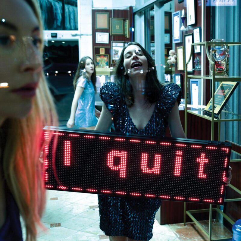 Fotografia em formato quadrado. No centro, Danielle Haim aparece com um vestido azul de paetês, segurando uma placa eletrônica vermelha com a frase “I quit” em letras minúsculas. Ela está atrás de um vidro, com reflexos de luz e objetos ao redor. Ao fundo, as irmãs Alana e Este observam a cena em segundo plano. O ambiente é uma loja com paredes envidraçadas, diplomas e prêmios expostos em prateleiras.