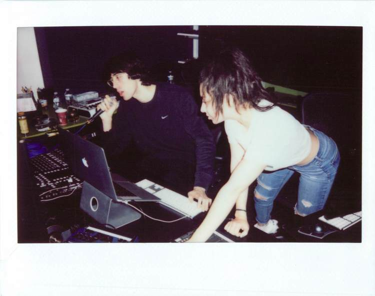 Uma foto Polaroid da Charli XCX e A. G Cook no estúdio. Ele segurando um microfone, enquanto olha para a tela de um laptop, a cantora está inclinada na mesa, e também olhando para a tela. A mesa está com aparelhos de produção de música.