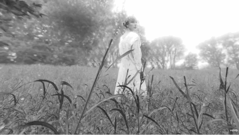 Imagem em preto e branco mostrando um campo extenso de vegetação alta, com árvores ao fundo suavemente embaçadas pela luz intensa. Taylor Swift aparece no centro da composição, de perfil, olhando por cima do ombro direito. Ela usa um vestido longo, claro e esvoaçante, com mangas bufantes e detalhes delicados, em um estilo romântico e campestre. Seus cabelos estão presos em um coque baixo e solto, e a luz cria uma aura difusa ao seu redor, quase etérea. A imagem evoca uma atmosfera de sonho, contemplação e afastamento.