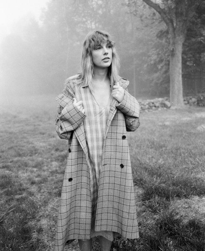 Foto em preto e branco de um campo gramado com árvores ao fundo, parcialmente ocultas pela névoa. Taylor Swift está em pé no centro da imagem, usando um vestido leve de tecido xadrez com decote em “V”, por baixo de um longo casaco também xadrez. Seu cabelo está solto, com franjas e ondas suaves, compondo um visual natural e descomplicado. Ela segura as lapelas do casaco com ambas as mãos, enquanto seu olhar está voltado para o horizonte, com uma expressão calma e distante. A imagem evoca uma estética rural, silenciosa e nostálgica, com um ar de delicadeza melancólica.