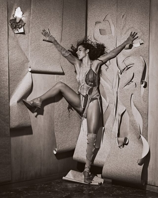 Fotografia em preto e branco de Marina Sena, em parte do ensaio para o álbum Coisas Naturais. Ela está vestindo uma roupa detalhada e botas de cano alto, com os braços estendidos e uma perna dobrada, em um ambiente estilizado com papéis rasgados criando uma textura visual. 