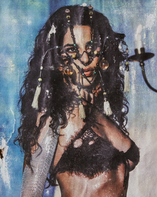 Close-up de Marina Sena com cabelos volumosos e um adereço facial e capilar intrincado, feito de tiras, contas e borlas. Ela veste um top preto texturizado e há um material prateado no braço esquerdo. O fundo é uma parede azul-acinzentada e desgastada, com um candelabro no canto superior direito. A foto tem uma estética granulada/vintage.