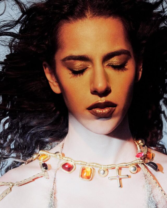 Close-up de Marina Sena com cabelos volumosos e um adereço facial e capilar intrincado, feito de tiras, contas e borlas. Ela veste um top preto texturizado e há um material prateado no braço esquerdo. O fundo é uma parede azul-acinzentada e desgastada, com um candelabro no canto superior direito. A foto tem uma estética granulada/vintage.