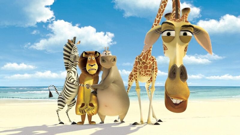 Cena do filme Madagascar. Na imagem, os quatro personagens principais — Alex, o leão; Marty, a zebra; Melman, a girafa; e Gloria, a hipopótamo — estão em uma praia tropical. Eles estão lado a lado, parecendo surpresos e confusos. Ao fundo, há um mar azul brilhante e céu limpo. A areia é clara, a iluminação é forte e ensolarada, transmitindo o clima de um ambiente quente. Os personagens estão posicionados de frente, encarando algo fora da imagem.