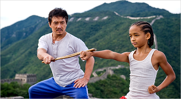 Fotografia de Jackie Chan, homem asiático de rugas, bigode e cavanhaque, vestindo calça azul da adidas e blusa branca de botões desgastada, e Jaden Smith, menino negro de tranças verticais, em uma regata branca, praticando movimentos de kung fu em meio às montanhas florestadas e a Muralha da China.