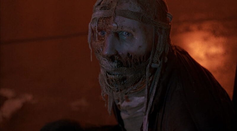 Cena do filme DarkmanNa imagem, o personagem Darkman olha para frente com expressão de desespero. Ele possui o rosto desfigurado, com cicatrizes na região da boca, bochechas e olhos, com exceção do lado esquerdo do rosto e o nariz. Na face, há ataduras. Darkman é um homem de pele branca e olhos azuis. Ele veste um paletó preto. 