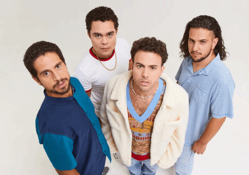 A imagem mostra os quatro integrantes, que posam em estúdio com fundo branco, olhares direcionados à câmera em uma leve perspectiva de cima para baixo. Um veste camisa azul com botões, outro camiseta branca com gola vermelha e colar dourado, o terceiro aparece com casaco felpudo branco sobre colete colorido, e o último usa camisa polo listrada azul e calça jeans. O visual do grupo combina elementos modernos com um toque retrô, transmitindo atitude e estilo despojado.