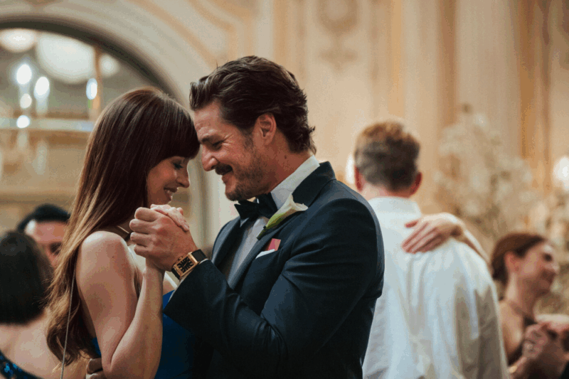 Cena do filme Amores Materialistas. A cena mostra Pedro Pascal e Dakota Johnson dançando de mãos dadas em um ambiente festivo, casamento. O foco está no casal, com o fundo desfocado, reforçando a intimidade do momento. O homem, de terno escuro, e a mulher, com um vestido azul, exibem expressões gentis e afetuosas. A iluminação suave e o uso da profundidade de campo criam um tom romântico e caloroso, transmitindo a atmosfera alegre e acolhedora do evento.