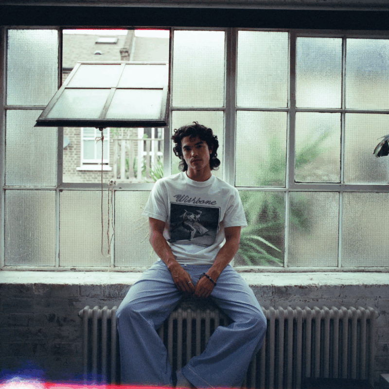 Pessoa sentada em um peitoril de janela grande com vidro fosco. Veste camiseta branca estampada com a palavra “Wishbone” e calça azul larga. O ambiente é iluminado de forma suave, criando um clima intimista e urbano.