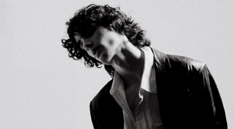 Uma foto em preto e branco do artista Sombr. O jovem, com cabelo escuro e encaracolado, está com a cabeça inclinada para baixo e para o lado. Ele usa uma jaqueta escura sobre uma camisa clara. A iluminação dramática cria um forte contraste e sombras em seu rosto. O fundo é liso e claro.