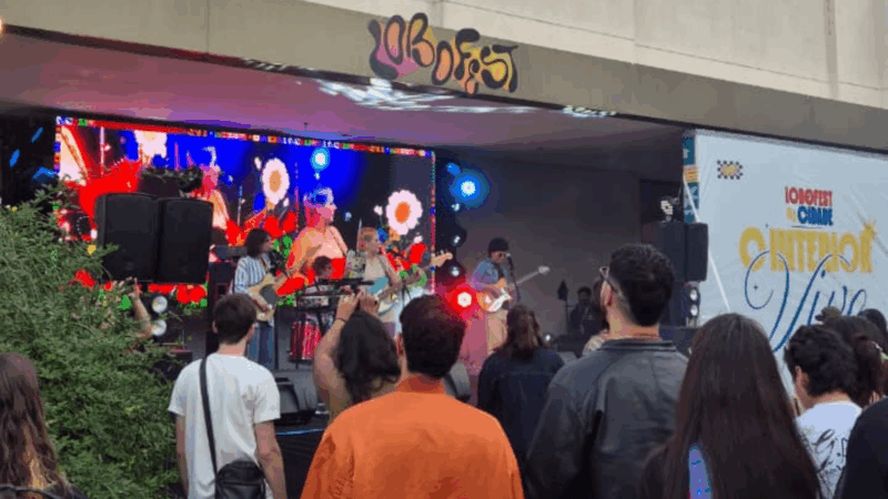 Uma foto de uma banda se apresentando em um palco no festival Lobofest em Sorocaba. O palco tem um grande telão de LED no fundo exibindo gráficos coloridos, incluindo flores. No primeiro plano, uma multidão de pessoas, vistas de costas, assistem ao show. Acima do palco, o nome "Lobofest" é visível. À direita, um grande banner vertical exibe os dizeres "Lobofest na Cidade / O INTERIOR VIVE".