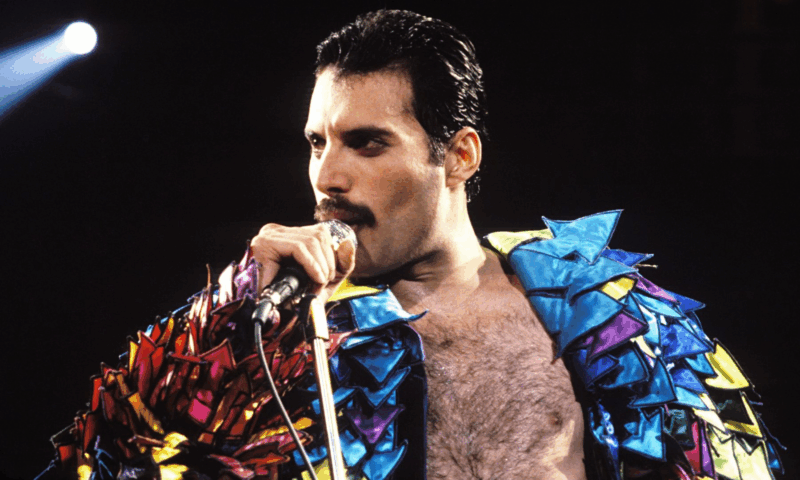Foto do artista Freddie Mercury cantando no palco com expressão intensa, vestindo uma jaqueta brilhante cheia de franjas coloridas. Ele segura o microfone com as duas mãos e está em destaque sob a luz de palco, com o peito parcialmente exposto e cabelo penteado para trás.