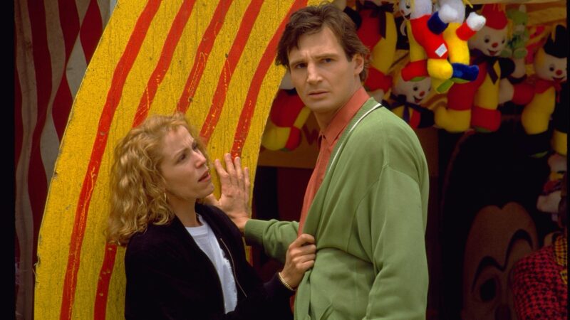  Cena do filme DarkmanNa imagem, a personagem Julie (à esquerda) e Peyton (à direita) estão em um parque de diversões. Ela segura a blusa dele, na região da barriga, e está com expressão de mágoa enquanto conversa com Peyton. Ele olha para a frente com expressão de dúvida. Julie é uma mulher branca, de cabelos loiros e ondulados, de altura baixa, na faixa dos 30 anos. Ela veste uma blusa de frio na cor preta, com o zíper aberto, revelando uma camiseta branca por baixo. Peyton é um homem branco, de cabelos lisos na cor castanho claro. Ele é alto e veste uma blusa de frio verde e uma camisa de botões na cor laranja. Ele apoia a mão direita em um dos estandes do parque de diversão. A estrutura é na cor amarela e possui riscos vermelhos, assim como sinais de desgaste, como arranhões e rachaduras. Ao fundo, pendurado no teto, há bonecos de palhaços de pelúcia. 