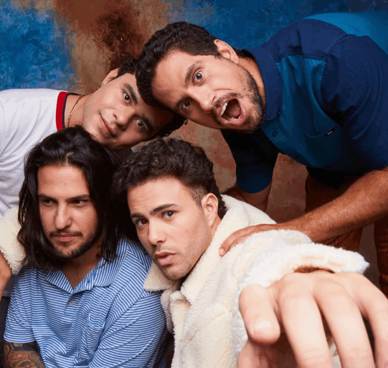 A imagem mostra quatro homens posam juntos para uma foto em um cenário colorido com fundo azul e marrom. Dois deles estão na parte superior da imagem, inclinando-se para frente — o da esquerda veste uma camiseta branca com detalhes vermelhos e o da direita, uma camisa azul, com expressão animada e boca aberta. Na parte inferior, dois outros homens estão mais próximos da câmera — um com cabelo comprido e camisa listrada azul, o outro com cabelo curto, expressão séria e vestindo um casaco branco felpudo, estendendo o braço em direção à câmera.