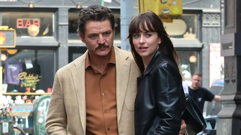 Cena do filme Amores Materialistas. A cena retrata um casal, Pedro Pascal e Dakota Johnson, em um ambiente urbano, com estilo casual e foco nas expressões e interações sutis entre eles. O homem, mais alto, tem cabelos castanho-escuros, bigode e veste casaco bege com camisa castanho-alaranjada, exibindo expressão neutra. A mulher, de cabelos castanho-escuros em camadas com franja, veste um casaco de couro preto e apresenta semblante levemente sério. O enquadramento é próximo, destacando o casal contra um fundo urbano desfocado, criando profundidade e realce visual. A iluminação natural e difusa indica uma cena ao ar livre durante o dia, com tons neutros e abordagem realista, sem efeitos estilísticos marcantes.