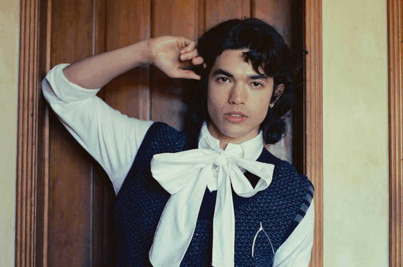 Pessoa de cabelos escuros e cacheados posa encostada a uma porta de madeira. Usa camisa branca com grande laço no pescoço e colete azul escuro texturizado. O olhar é direto para a câmera, transmitindo intensidade.