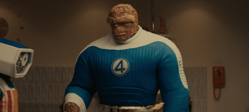 A imagem mostra o Coisa, com uma roupa azul e branca, em um ambiente doméstico. O fundo da imagem é composto por uma parede de cor neutra e um telefone vermelho de botão. A iluminação parece suave, sem fortes contrastes. Coisa é a figura principal, com pele de cor marrom-terra e um visual robusto. O seu traje é azul e branco com detalhes que lembram roupas dos anos 60/70. O ambiente parece ser um apartamento ou uma cozinha, com elementos de decoração clássicos de décadas anteriores. A iluminação é natural e homogênea, sem sombras acentuadas e a cor de fundo é um tom suave e quase monocromático.