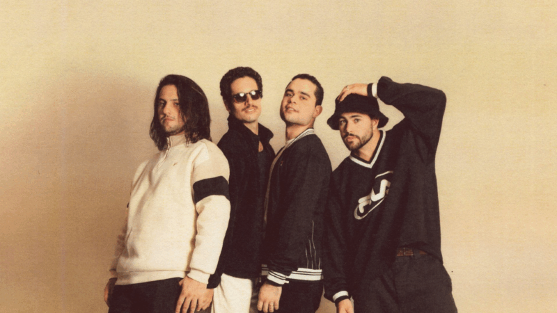 A imagem mostra quatro homens posam em frente a um fundo neutro bege. O grupo exibe um estilo casual e retrô: um veste moletom branco com faixa preta na manga e tem cabelo longo; outro usa óculos escuros e bigode, com jaqueta preta; o terceiro veste jaqueta esportiva e está centralizado, com expressão confiante; o último usa suéter preto com logotipo da Fila e chapéu tipo bucket, com uma mão na cabeça. A iluminação suave e o filtro quente evocam uma estética nostálgica dos anos 90.