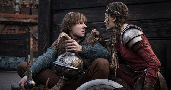 Cena do filme Como Treinar o Seu Dragão. Dois personagens estão sentados próximos a uma parede de madeira escura. À esquerda está o personagem Soluço, um jovem branco e com cabelos castanhos. Ele veste uma blusa de lã azul e um colete de marrom, e segura um elmo prateado com uma mão, enquanto apoia a outra em um machado. À direita está a personagem Astrid, uma jovem negra com seus cabelos trançados em dois rabos. Astrid usa uma armadura metálica nos ombros e uma túnica vermelha por baixo. Ela segura Soluço pela gola da blusa e o encara com firmeza 