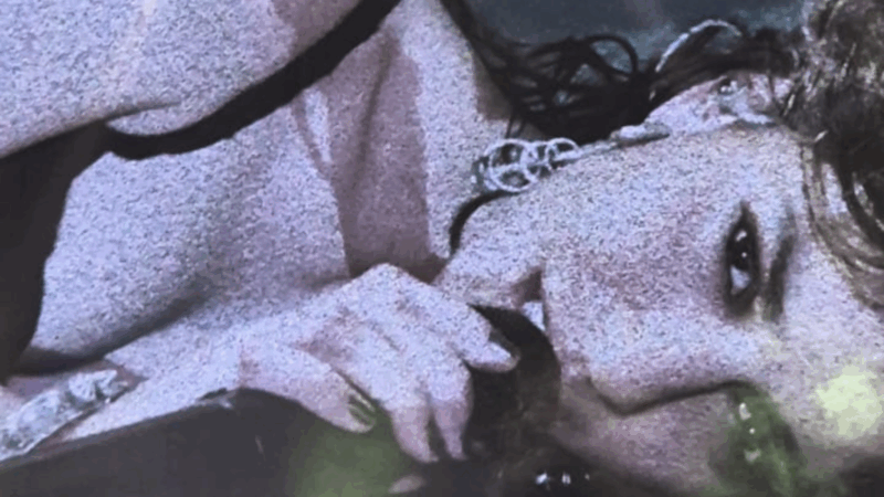 Uma foto granulada em close-up e com inclinação diagonal da cantora Marina Sena durante um show. Ela segura um microfone com a mão, com as unhas pintadas, e o leva em direção à boca. Seu cabelo escuro e encaracolado, parte do rosto e um de seus olhos, que olha diretamente para a câmera, são visíveis. Ela também usa um colar com um pingente circular.