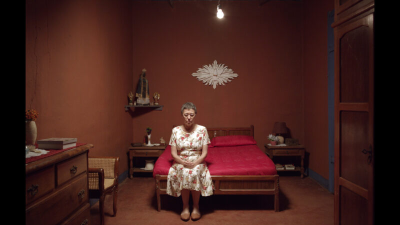 Imagem do filme Nada. Uma mulher de meia-idade, com cabelos curtos e grisalhos, veste um vestido claro com pequenas estampas florais e está sentada na beirada de uma cama coberta por um tecido vermelho intenso. Suas mãos estão repousadas no colo e seu olhar é fixo. Ela está no centro de um quarto com paredes pintadas em um tom terroso avermelhado, iluminado por uma única lâmpada pendente no teto, criando sombras suaves. À sua esquerda, sobre uma pequena prateleira fixada na parede, há uma imagem sacra, um pequeno frasco e um objeto não identificável. À direita da cama, uma porta de madeira escura está parcialmente visível. A composição da cena é fortemente simétrica, com a mulher no ponto focal. Um elemento decorativo branco em formato floral adorna a parede acima da cabeceira da cama.