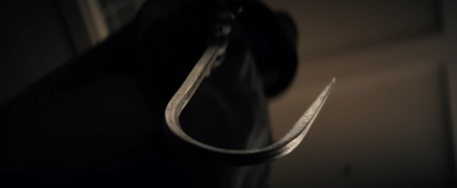 Cena do filme Eu Sei O Que Vocês Fizeram No Verão PassadoNa imagem, a câmera está no ângulo de baixo para cima, com o gancho do assassino em destaque. A arma possui formato de anzol e é prateado. O assassino está em desfoque, ele usa capa de chuva e chapéu, ambos na cor preta. 